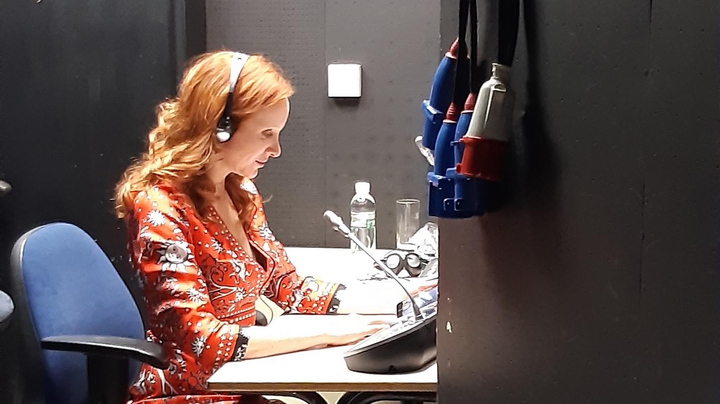 Zvočna opisovalka Mojca Mavec v rdeče-beli obleki sedi za mizo v komentirnici Cankarjevega doma. Gleda v list papirja. Pred njo je prenosni mikrofon. Foto: MMC RTV SLO/Veronika Rot Zvočna opisovalka Mojca Mavec v rdeče-beli obleki sedi za mizo v komentirnici Cankarjevega doma. Gleda v list papirja. Pred njo je prenosni mikrofon. Foto: MMC RTV SLO/Veronika Rot